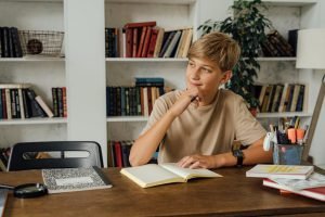 A thoughtful teenager pondering at a study desk surrounded by bookshelves, holding a pen.