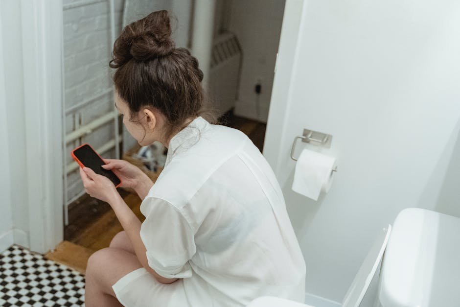 A woman wearing a white shirt uses a smartphone while sitting in a modern bathroom, suggesting relaxation and comfort.