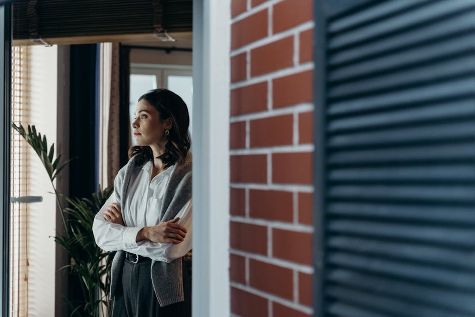 A contemplative woman stands indoors, arms crossed, gazing out the window beside a brick wall.