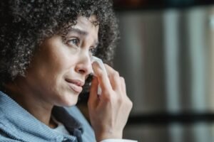 African American woman with curly hair wipes tears with tissue in a quiet indoor setting.