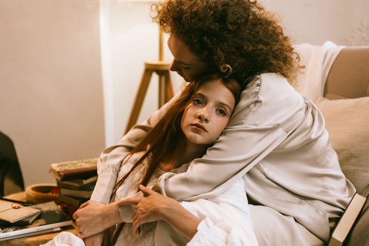 A woman lovingly embraces a teenage girl in a warm home interior.