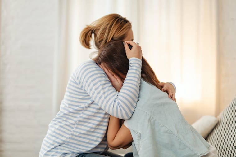 A mother lovingly comforts her teenage daughter in an emotional embrace indoors.