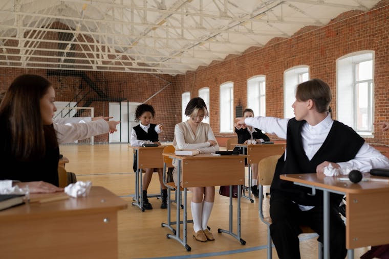 Teenagers in school uniforms pointing at classmate indoors, highlighting bullying.