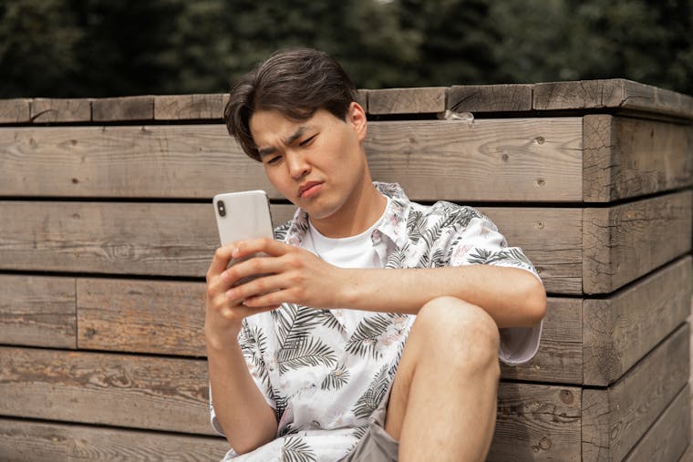 Young ethnic male in stylish clothes leaning on wooden barrier while messaging on mobile phone in park