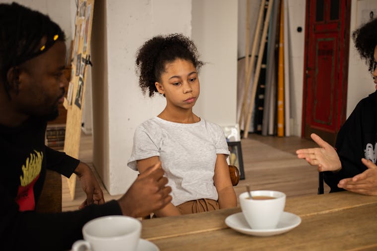 Sad African American girl sitting at wooden table with cups of coffee near black parents having conversation in light room