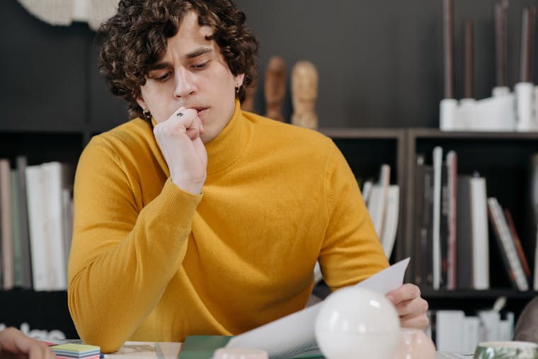 A thoughtful man in a yellow sweater closely examines a paper, seated indoors with books in the background.