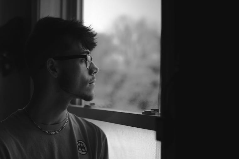 Black and white portrait of a man in eyeglasses looking out a window, feeling pensive.