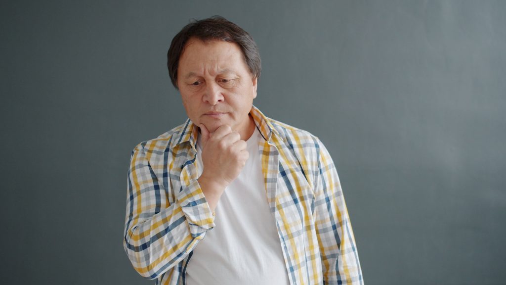 Thoughtful adult man in plaid shirt contemplating indoors against gray backdrop.