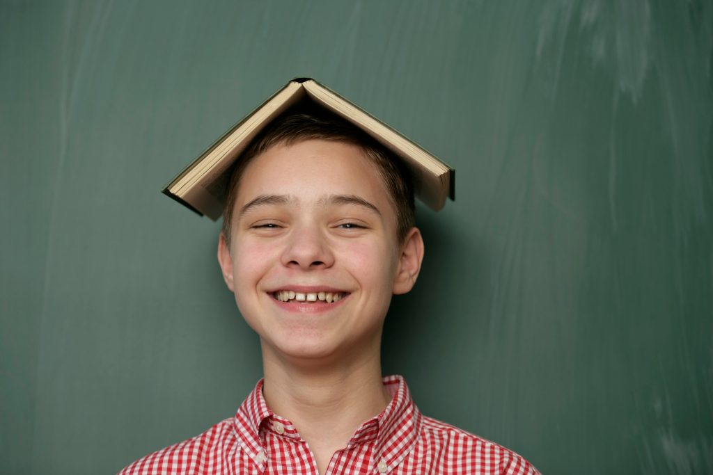 Cheerful teenager posing with book on head against chalkboard background.