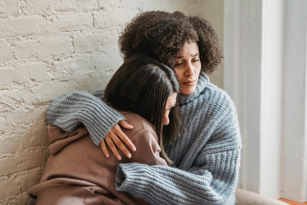Unhappy African American woman in casual wear hugging and consoling female friend while sitting near wall in room at home